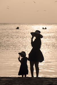 A mother holds her daughter's hand in front of the sea watching the sunset
