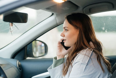 Beautiful girl with long hair in a grey trench coat using smartphone call gets into the car