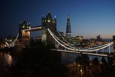 Illuminated bridge over river by buildings against sky at night