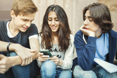 Happy girl using smart phone while sitting with male friends on steps