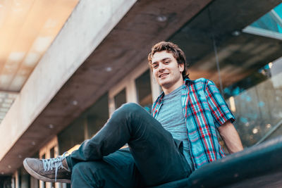 Low angle portrait of man smiling while sitting on seat against building