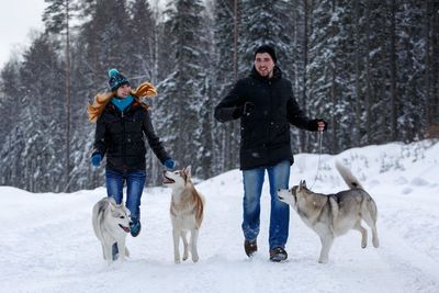 Young couple walking with siberian huskies on snowy field against trees