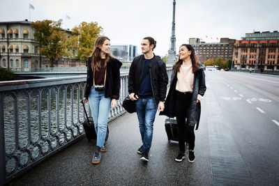 Male and female friends talking while walking with luggage on bridge in city