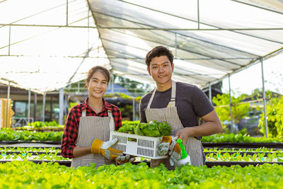 Smiling man and woman holding basket while standing in greenhouse