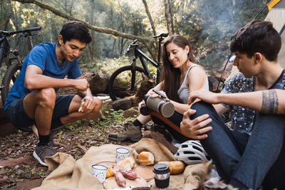 Diverse cyclists sitting on ground near tent and having picnic while hiking together in forest