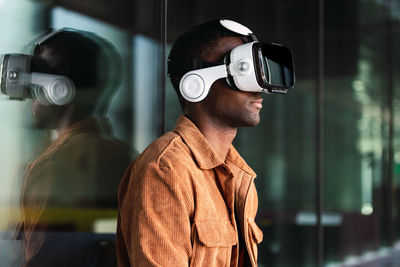 Positive african american male in modern vr goggles standing near glass wall while exploring virtual reality on street of city