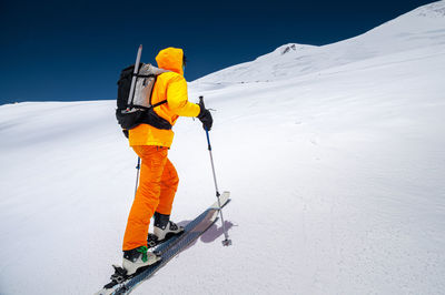 Young male freerider guide in a yellow ski suit with a backpack on his shoulders climbing 