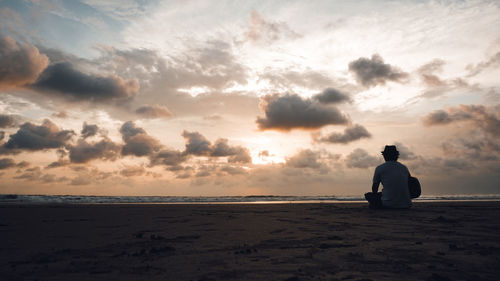 Rear view of man sitting on beach against sky during sunset