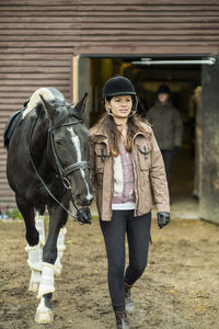 Young woman with horse walking outside barn with man in background