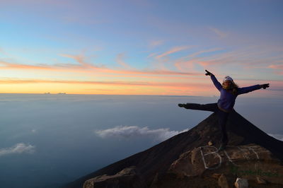Woman with arms outstretched standing on cliff against sky during sunset