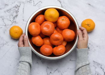 Female hands hold an iron bowl with ripe tangerines on a white table, overhead view