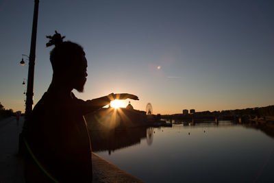 Man gesturing while standing by lake and city against clear sky during sunset