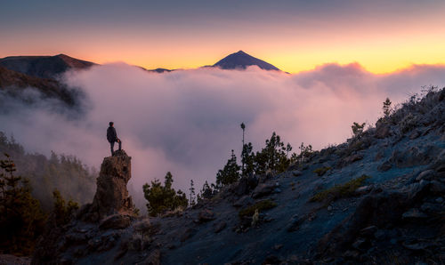 Back view of anonymous explorer standing on rock and observing majestic scenery of mountains under thick fog in morning during vacation in tenerife