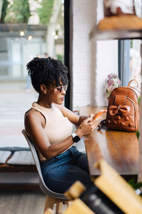 Side view of confident young african american female millennial with dark curly hair in trendy outfit and sunglasses sitting at table in modern cafe and messaging on smartphone