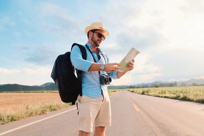 Man wearing hat holding map standing on road against sky