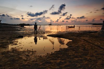 Silhouette man standing by camera and tripod at beach against sky during sunset