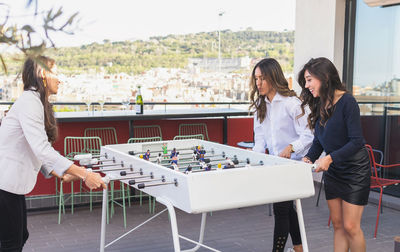 Happy adult hispanic girlfriends in smart casual clothes smiling and playing table football together during party on weekend day on terrace