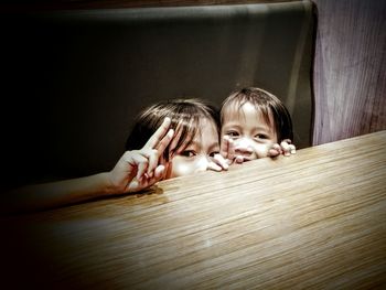 Sisters gesturing peace sign while hiding behind wooden table at home