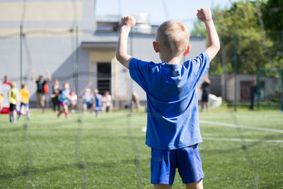 Rear view of a boy with arms raised looking through fence at playing field