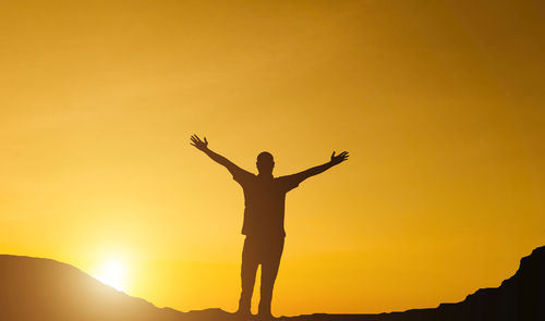 Silhouette man with arms outstretched standing against orange sky