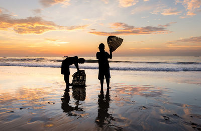 Silhouette boys holding birdcage while standing at beach against sky during sunset