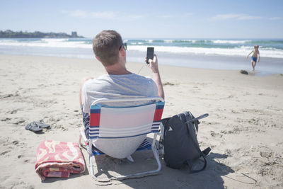 Rear view of man using mobile phone while sitting on chair at beach