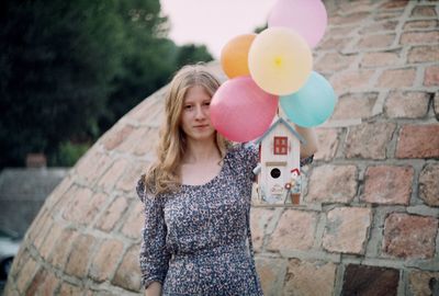 Portrait of young woman with balloons and model house standing against brick wall