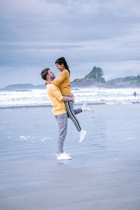Side view of man carrying woman while standing at beach against sky