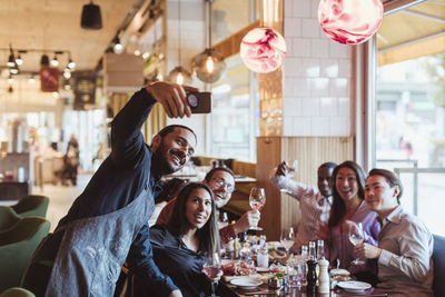 Happy waiter taking selfie on smart phone with customers in restaurant