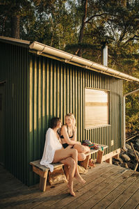 Female friends sitting on bench near cottage during sunset