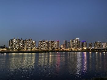 Illuminated buildings by river against clear sky at night