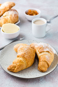 A pair of fresh crispy croissants on a plate, a cup of coffee and bowls of jam and butter 