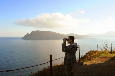 Man in gray military jacket looks  binoculars on high altitude mountain surrounded by sea, mountains 