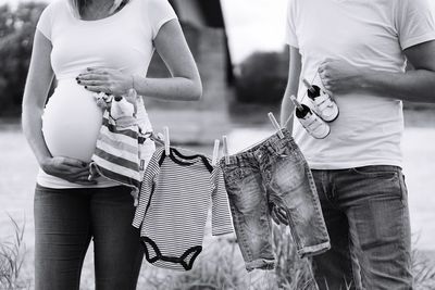 Midsection of pregnant woman and man holding baby clothing on field