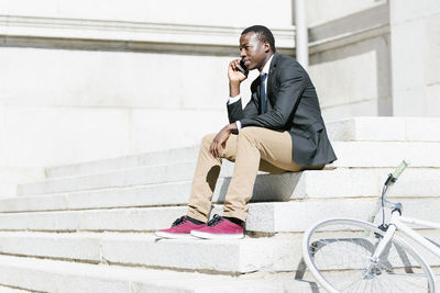 Businessman with bicycle talking on smart phone while sitting on steps in city