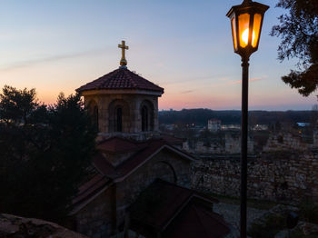 Illuminated street light by building against sky during sunset