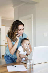Mother talking on phone while carrying baby boy looking at laptop computer
