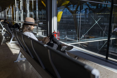 The guy in the hat at the airport in the waiting room sitting waiting for his flight, with wireless headphones to listen to music while chatting with his smart phone, side view