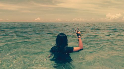 Rear view of woman in sea showing peace sign against sky