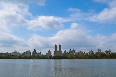 Scenic view of lake at central park by cityscape against cloudy sky