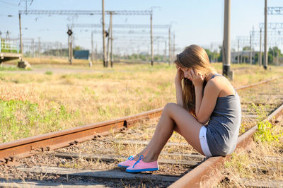 Depressed woman using phone while sitting on railroad tracks during sunny day