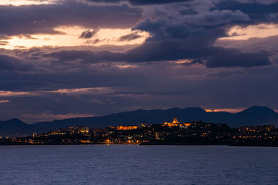 Sea by illuminated city against sky during sunset