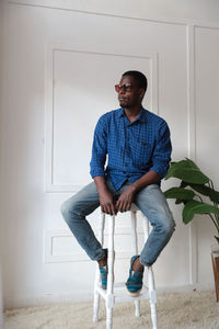 Full length of young man sitting on stool against wall