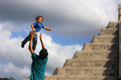 Rear view of man tossing cheerful son against kukulkan pyramid