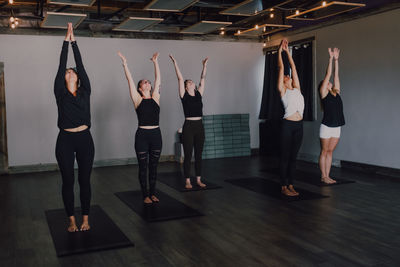 High angle of calm slender ladies in activewear with namaste standing in mountain position on sports mats on wooden floor and meditating in spacious contemporary gym looking up