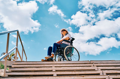 Low angle full body of positive handicapped female sitting in wheelchair near stairway looking away against blue sky in city