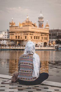 Rear view of woman sitting by lake and golden temple against sky