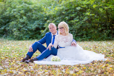 Portrait of smiling bride and groom sitting on field against plants at park