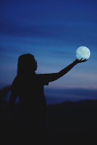 Optical illusion of silhouette woman holding moon against sky during dusk