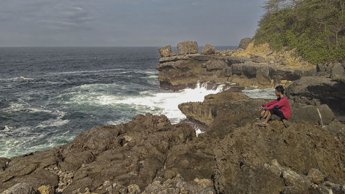 Rear view of boy standing on rock by sea against sky